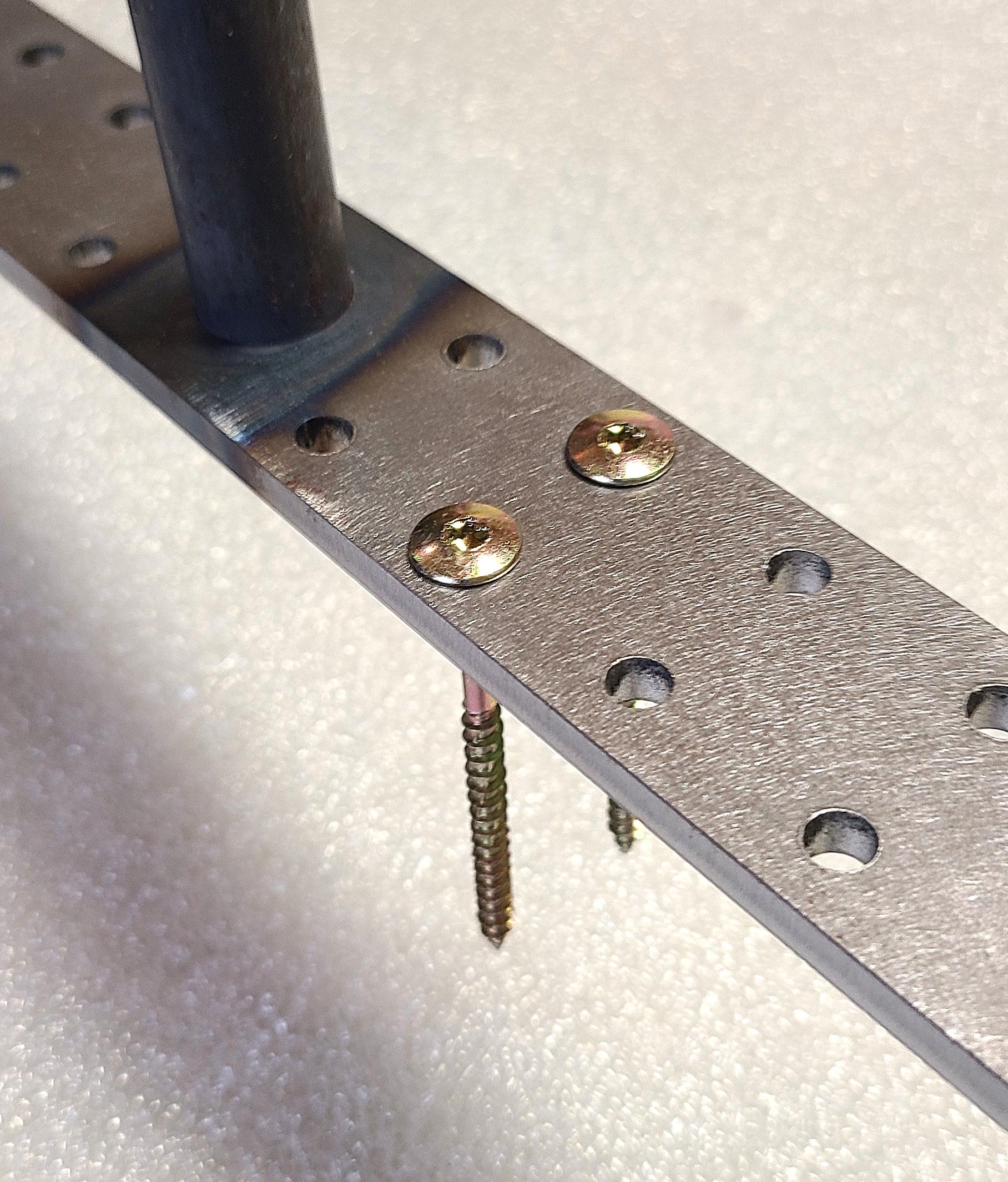 Close-up of a metal mounting bracket with pre-drilled holes, brass-colored screws, and a dark vertical rod attached, resting on a light textured surface.